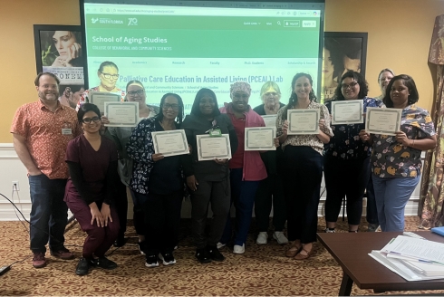 A group of palliative care trainers and assisted living staff stand together in a classroom holding certificates of completion. A presentation slide for the Palliative Care Education in Assisted Living Lab is displayed on a screen behind them.