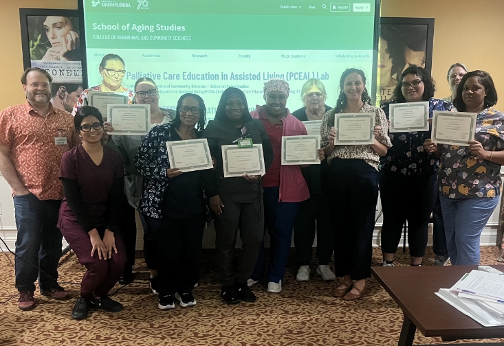 A group of palliative care trainers and assisted living staff stand together in a classroom holding certificates of completion. A presentation slide for the Palliative Care Education in Assisted Living Lab is displayed on a screen behind them.
