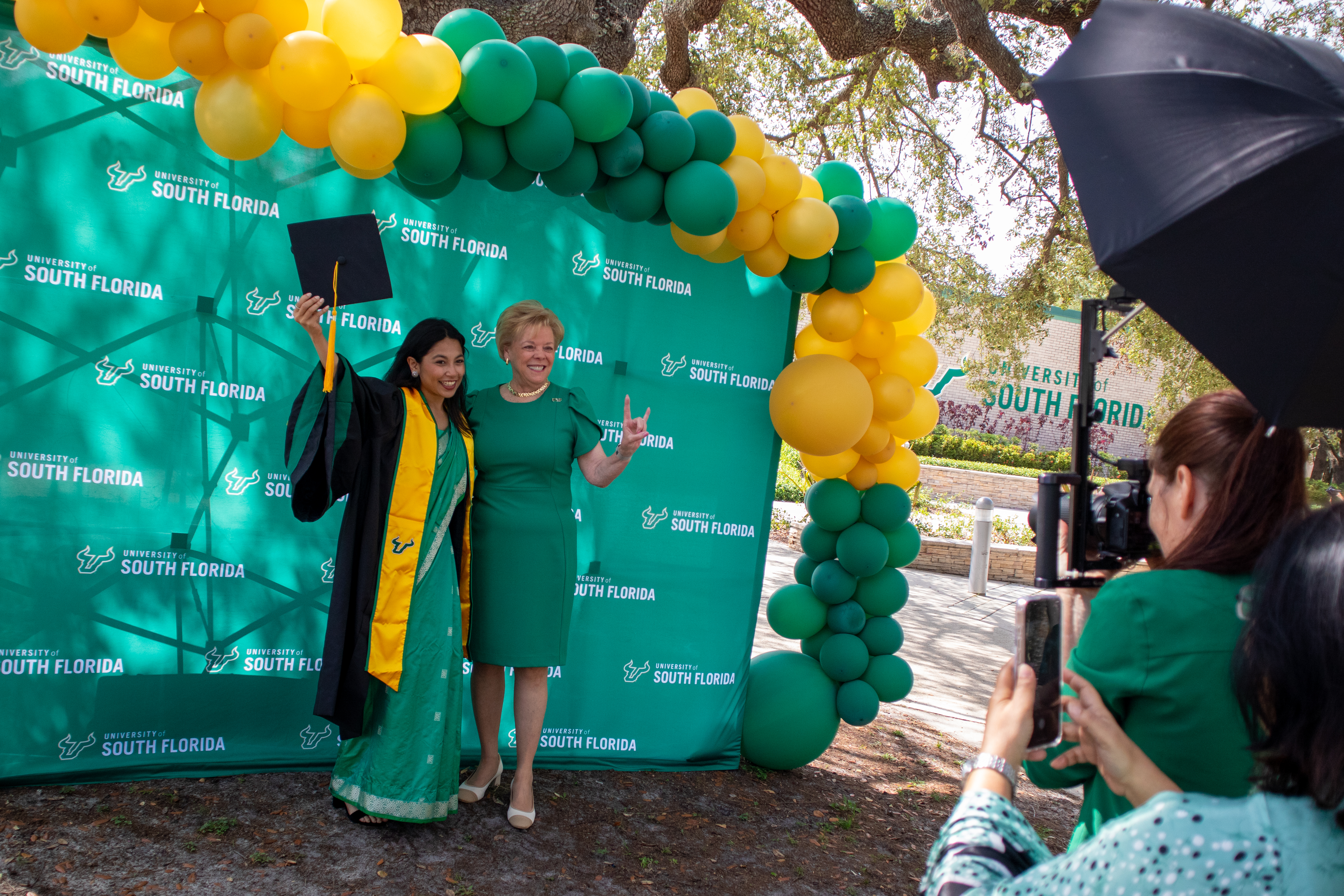 A graduating student stands beside USF President Rhea Law near green and gold balloons for a photo op