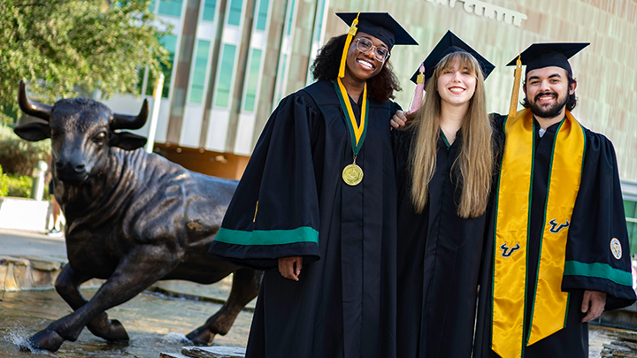 Two graduates of USF walk nearby a fountain in regalia and stoles.