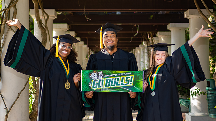 Three USF grads, wearing regalia, pose for the camera.