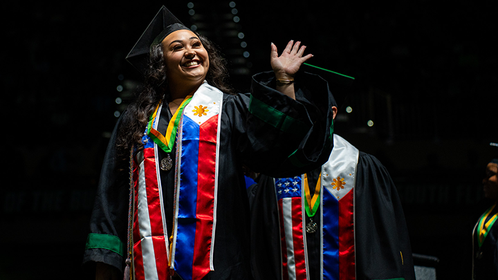 USF graduate wearing regalia walking across the Commencement stage waving to the crowd