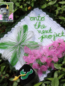 A fully knitted grad cap outfitted with a small, knit Rocky the Bull figure and a pink flower done in knitting yarn with knitted words that read "Onto the next project."
