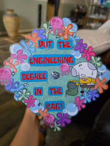 Grad cap decorated in bright blue, pink, and purple colors featuring the flowers from Spongebob's underwater world and cut out images of Spongebob and a jellyfish saying "Put the Engineering degree in the bag".