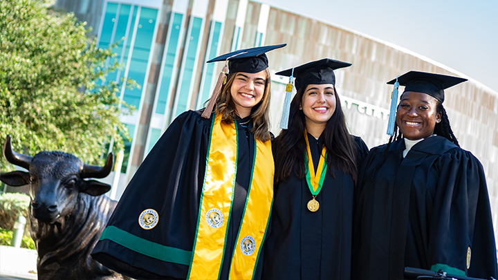 Three USF grads wearing regalia smile at the camera, standing in front of a Bull statue
