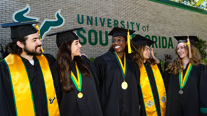Five USF grads, wearing regalia, smile at each other.