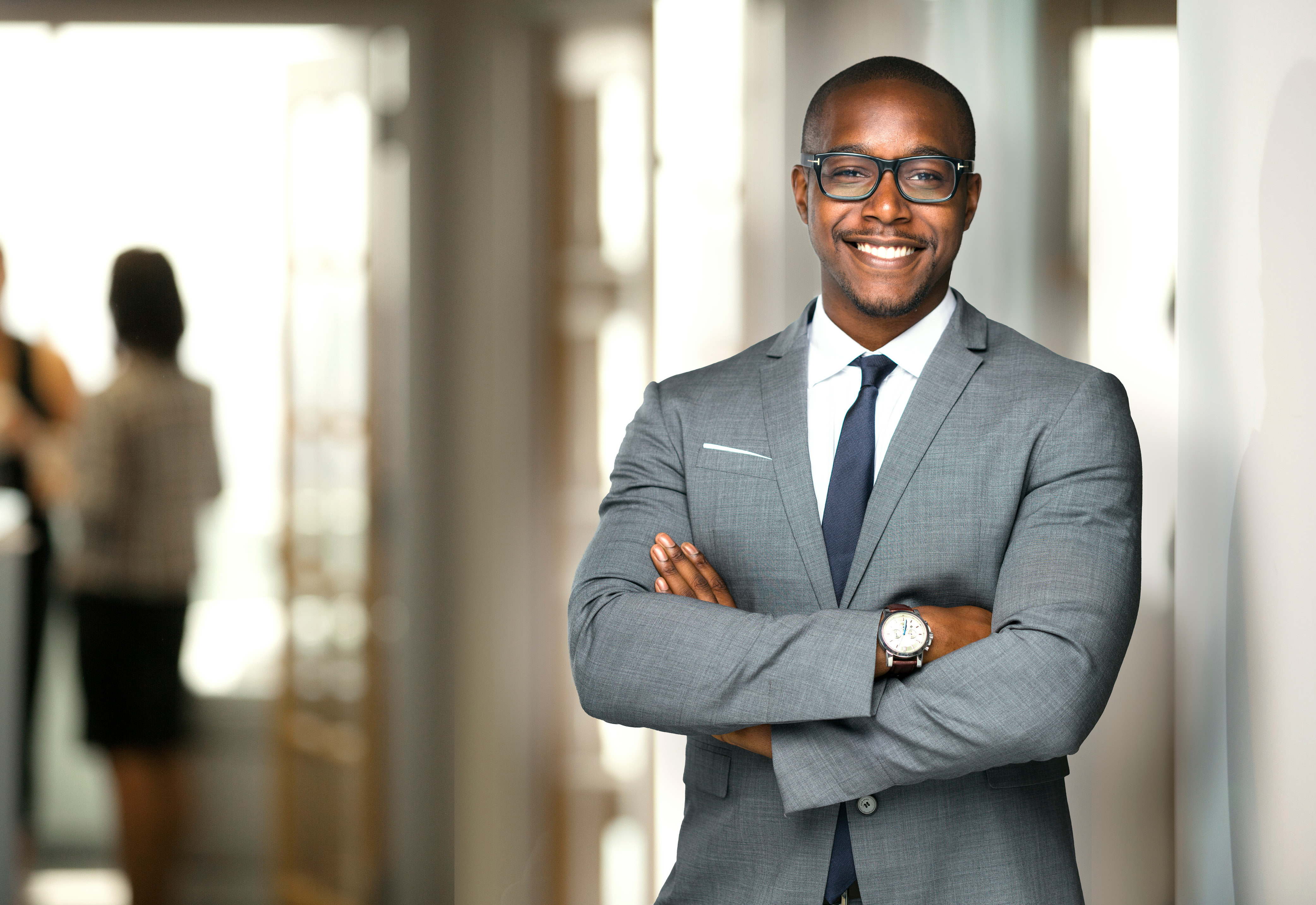 A man wearing a suit, glasses, and a watch standing with his arms crossed