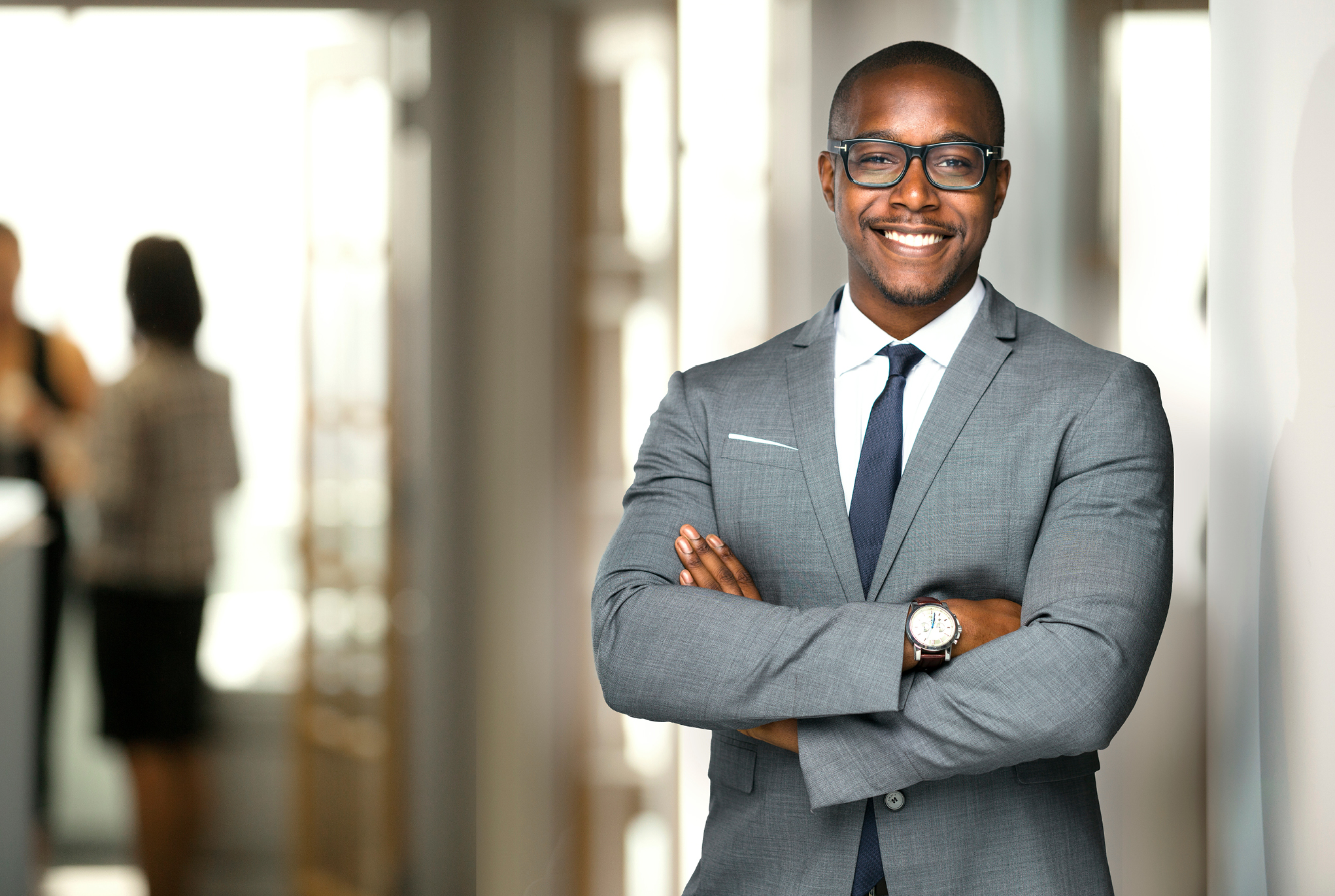 A man wearing a suit, glasses, and a watch standing with his arms crossed