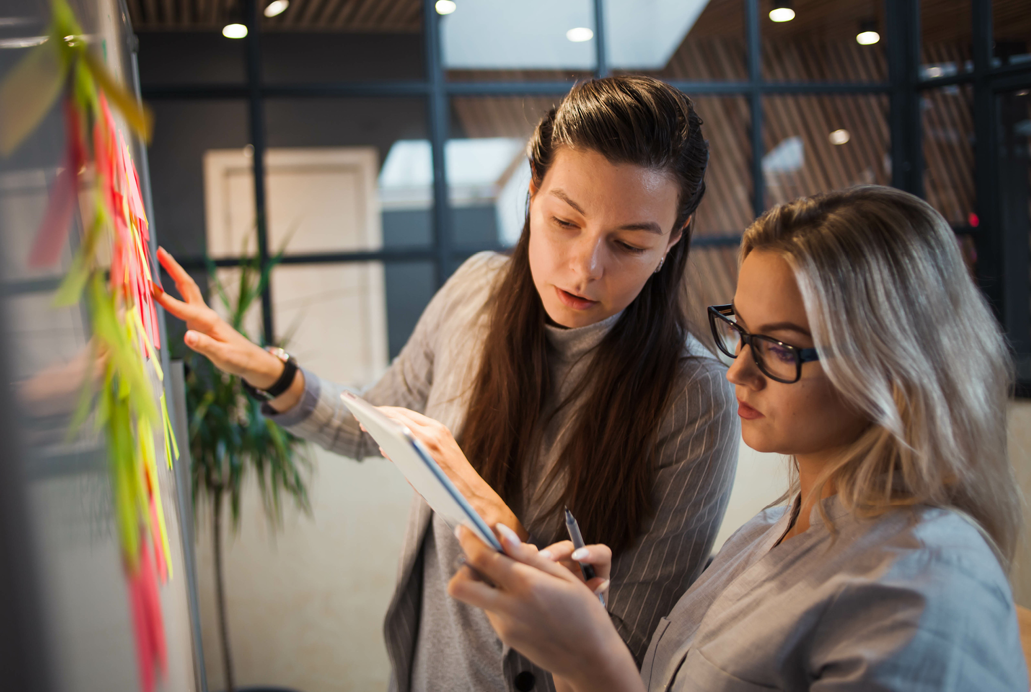 Two women looking at a tablet and standing in front of a board filled with sticky notes