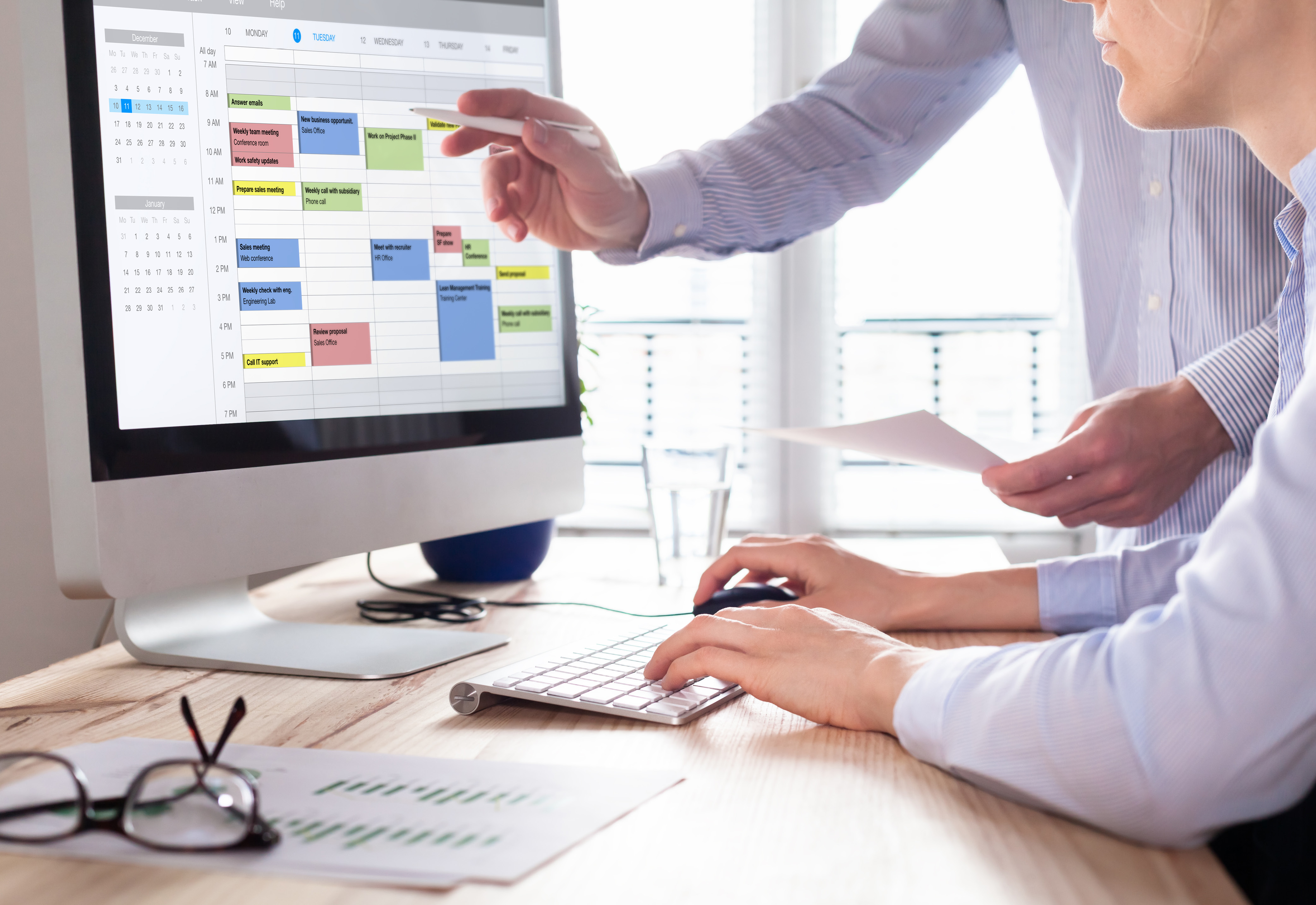 Two people at a desk with one pointing at a calendar on a computer screen and the other person typing