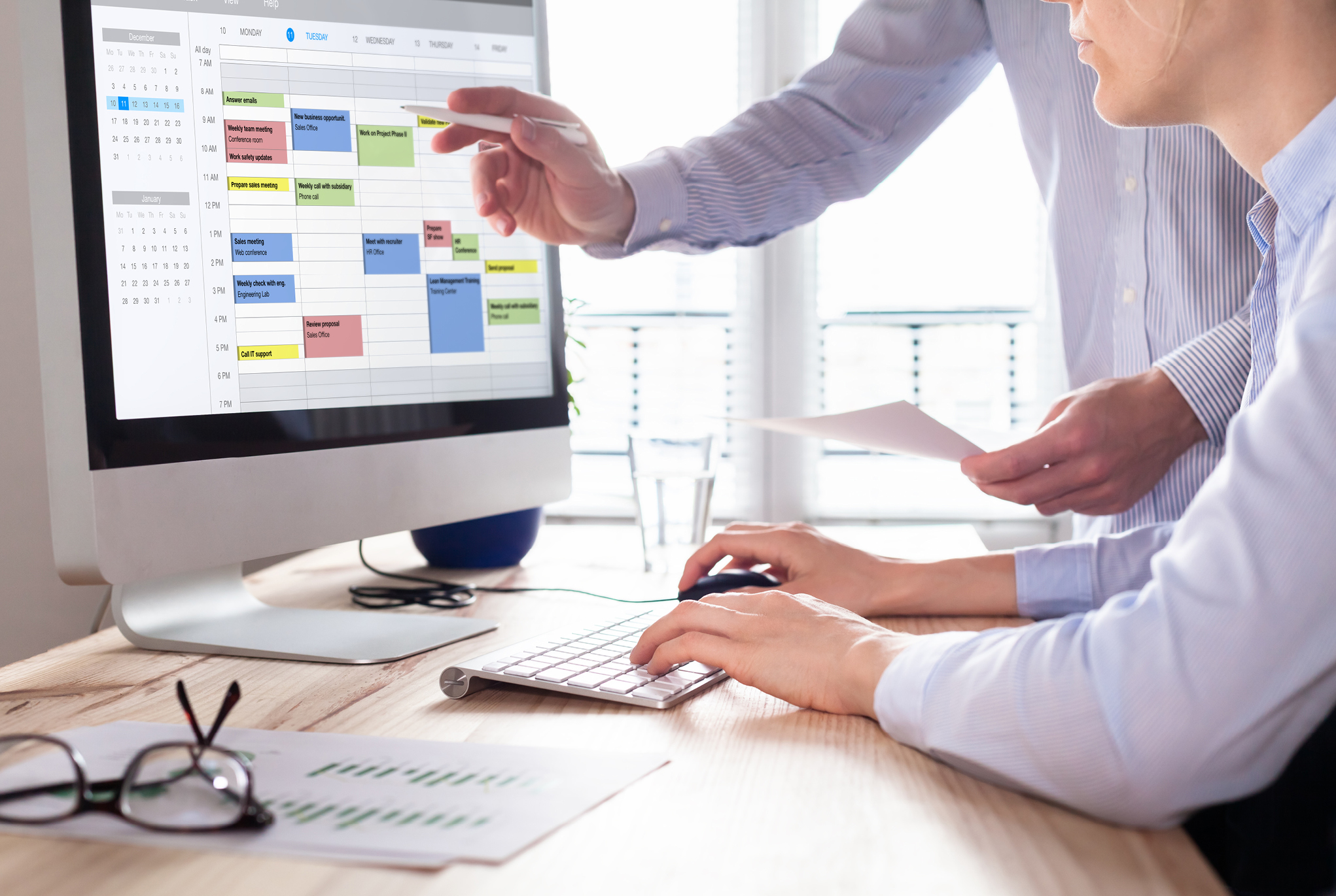 Two people at a desk with one pointing at a calendar on a computer screen and the other person typing