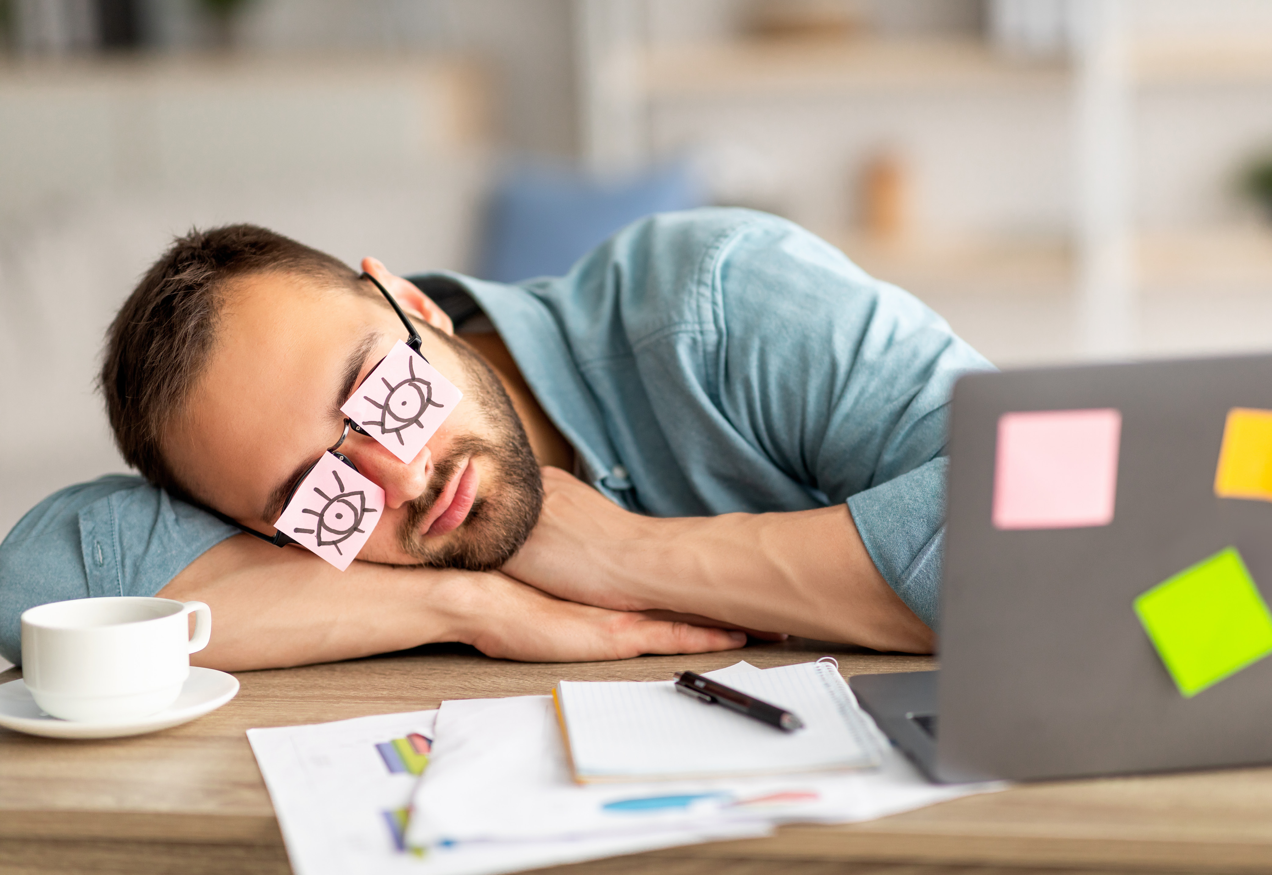 A man sleeping at his desk with eyes drawn on sticky notes on his glasses