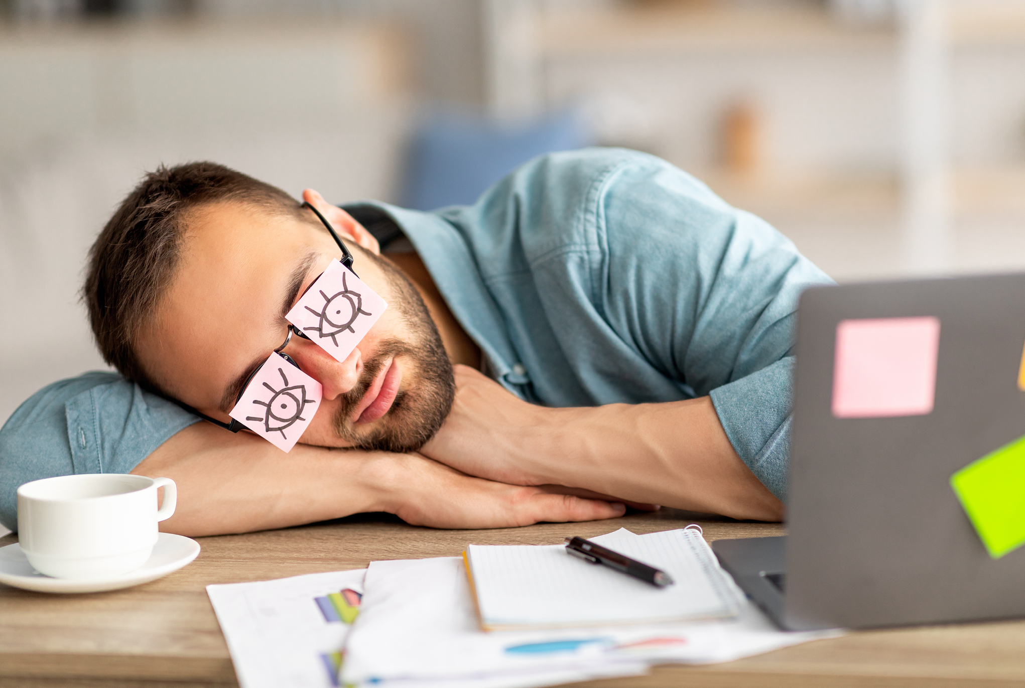 A man sleeping at his desk with eyes drawn on sticky notes on his glasses