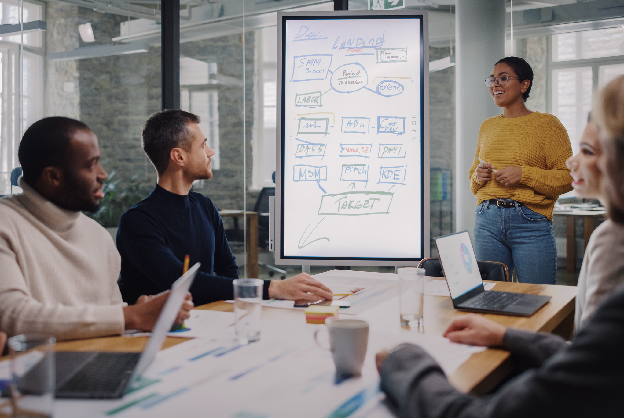 A group of people having a meeting while one person is giving a presentation on a white board. 