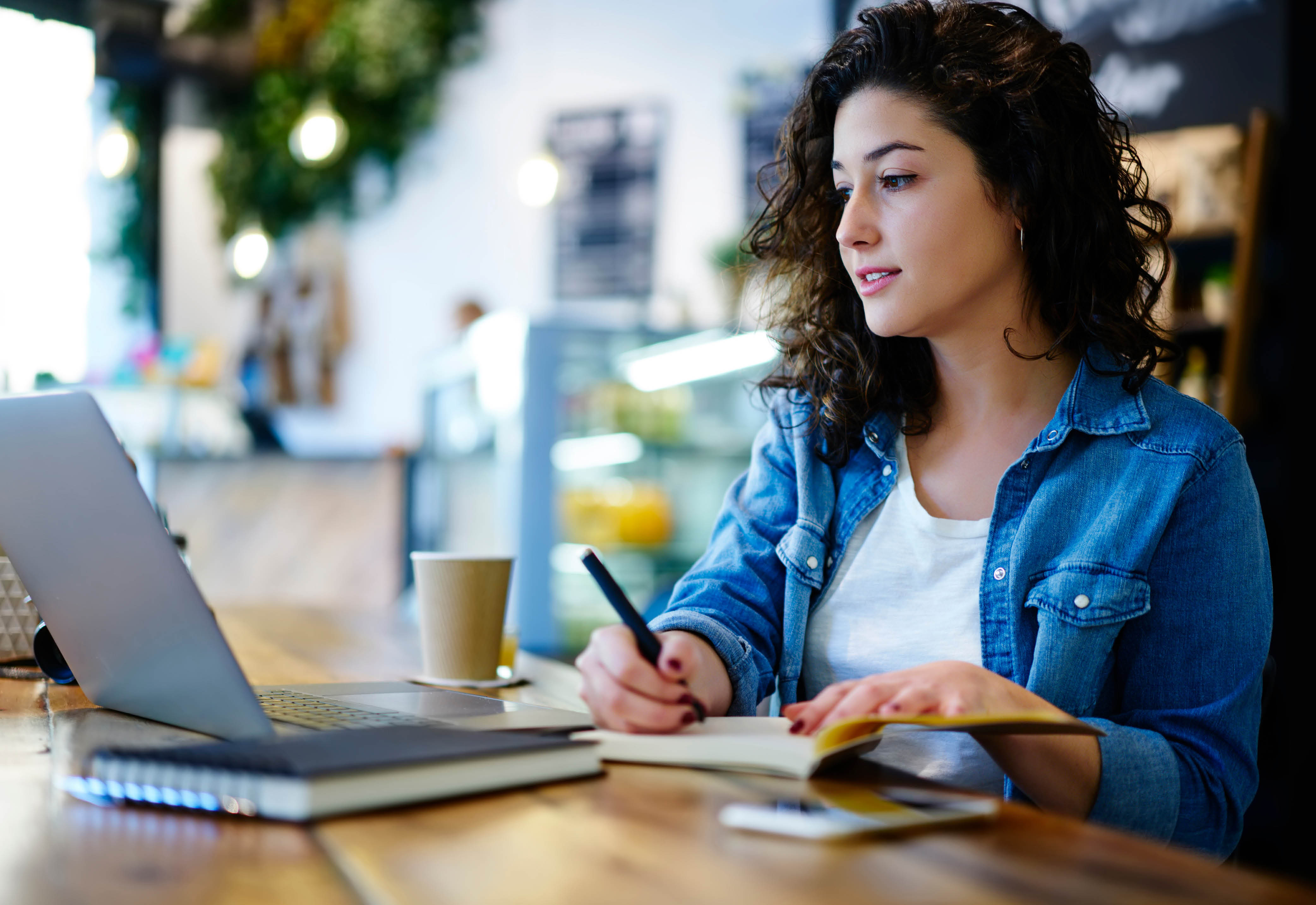 Woman in a white t-shirt and denim jacket looking at her laptop and taking notes