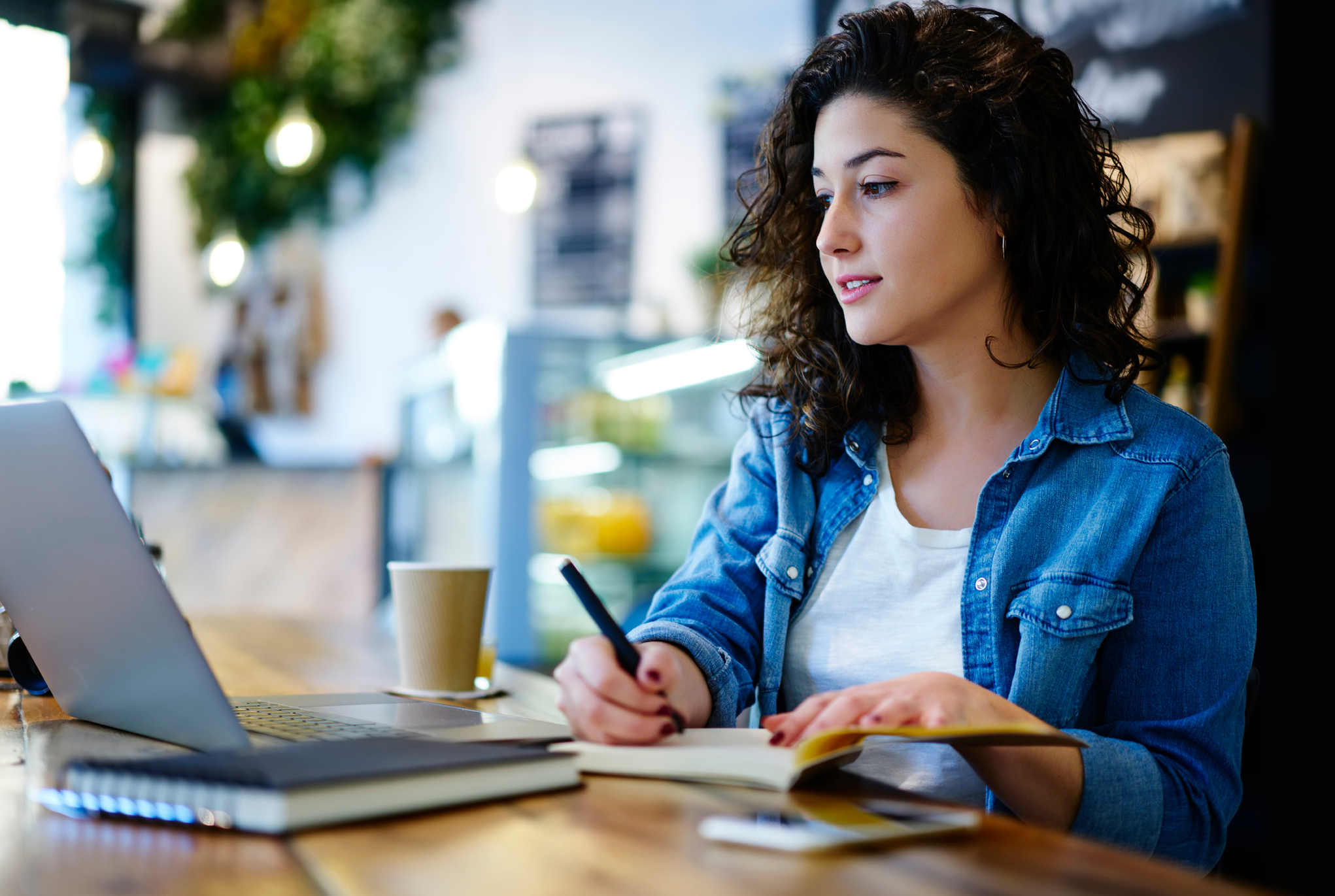 Woman in a white t-shirt and denim jacket looking at her laptop and taking notes