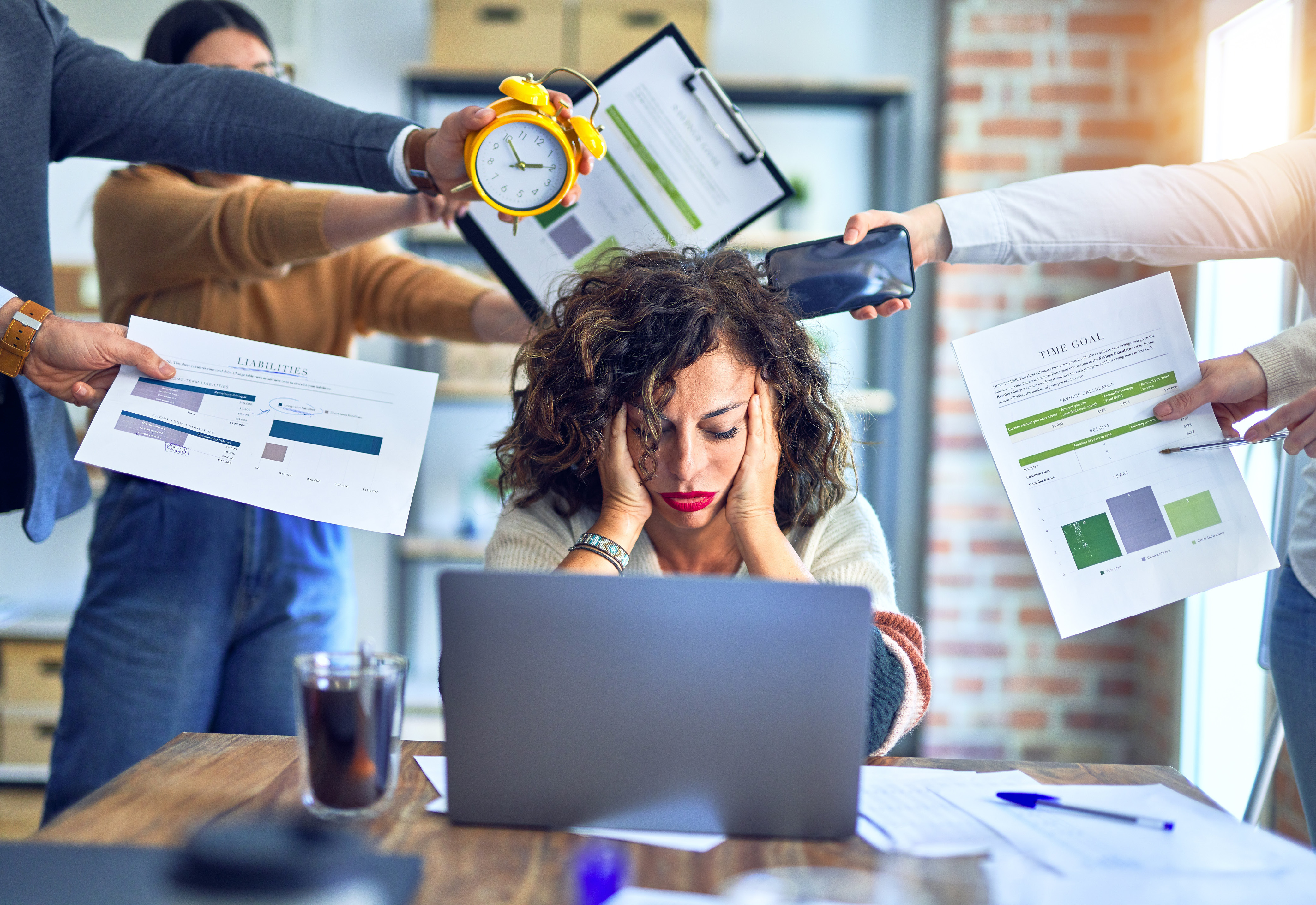 A woman sitting at her desk with her head in her hands and several people holding up stressful items around her