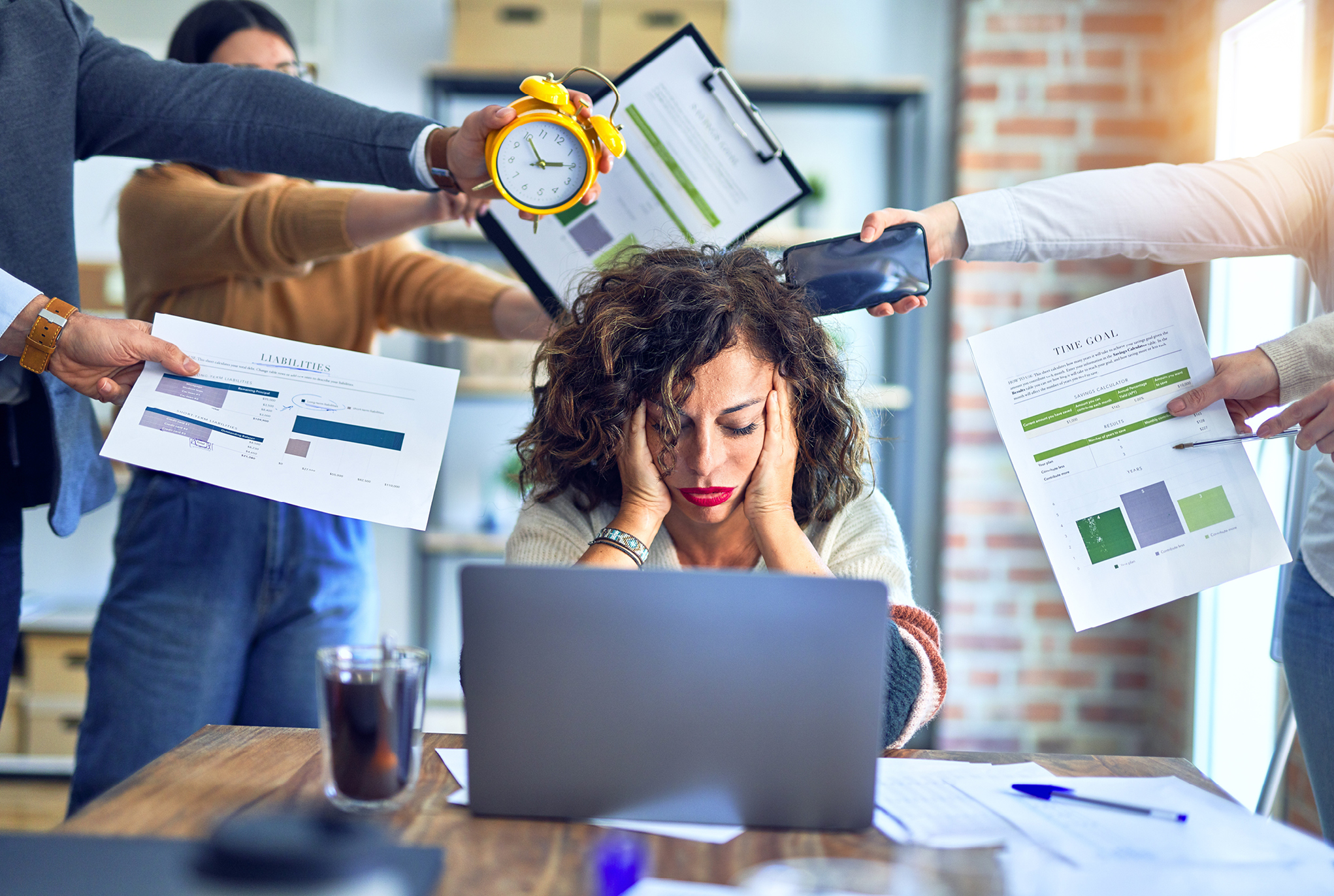 A woman sitting at her desk with her head in her hands and several people holding up stressful items around her