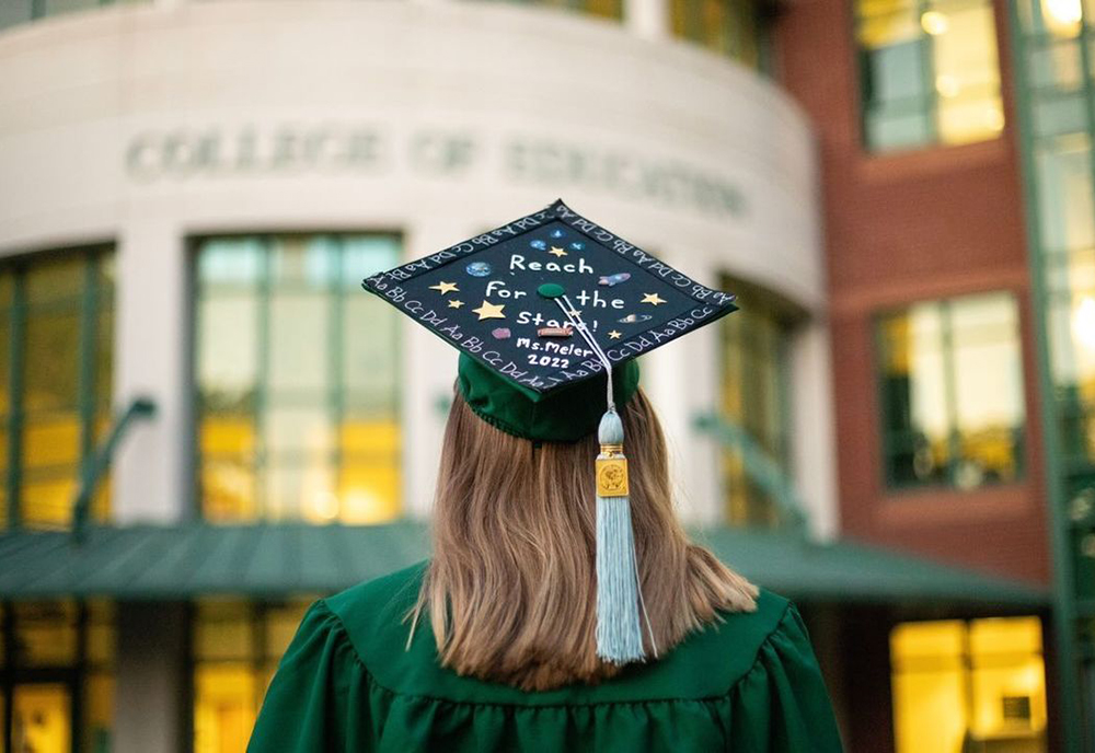USF graduate standing outside the College of Education building with decorated graduation cap