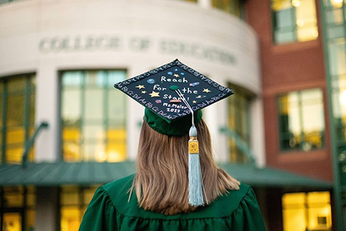 USF graduate standing outside the College of Education building with decorated graduation cap
