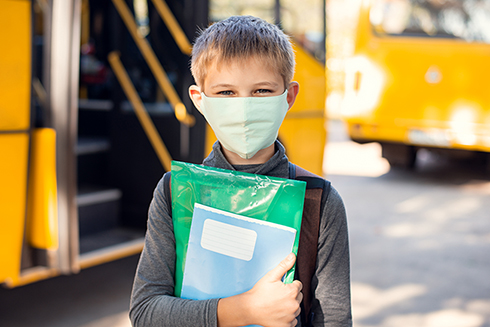 Small school boy in medical mask holding learning materials standing near a school bus