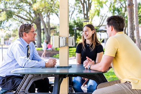 Two students and a professor talking outside at a table