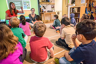 Classroom of children seated on the floor paying attention to teacher and presentation on screen