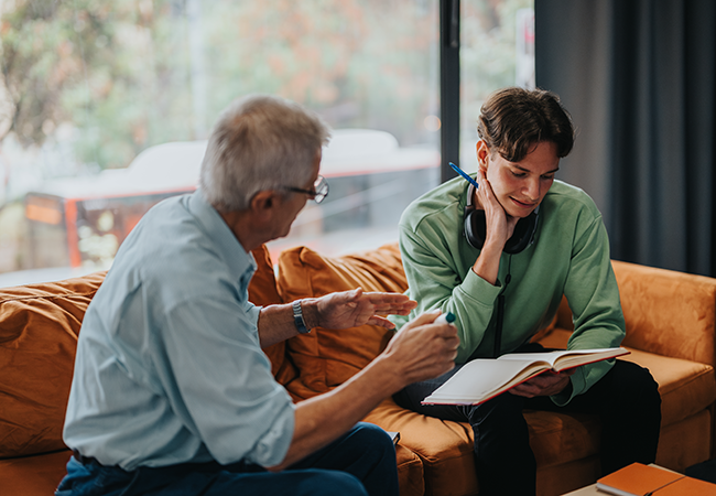 2 men sit on a couch while one talks to the other with a book in hand