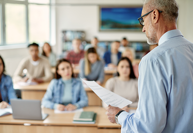 a male professor stands and teaches at the front of a classroom