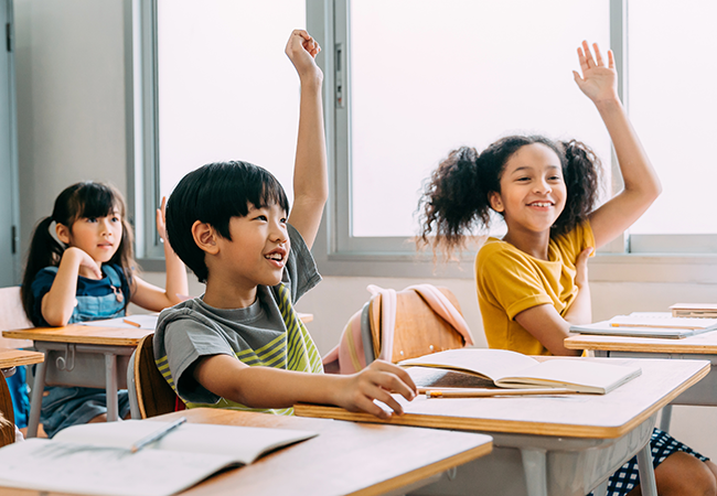 two young children seated in a classroom raising their hands