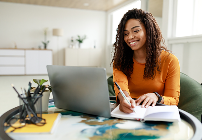 smiling woman takes notes in a notebook while seated at laptop