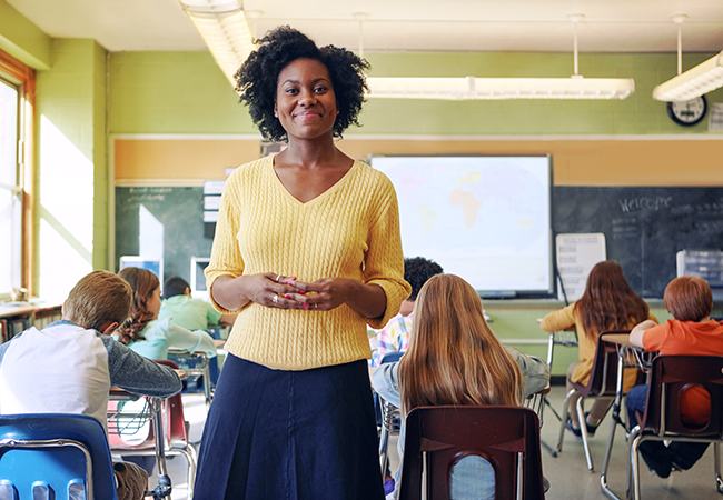 a teacher stands at the back of a classroom smiling at camera