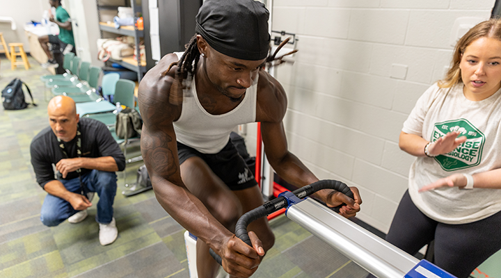 Athlete during the wingate test while a faculty member and an exercise science and kinesiology student watch on