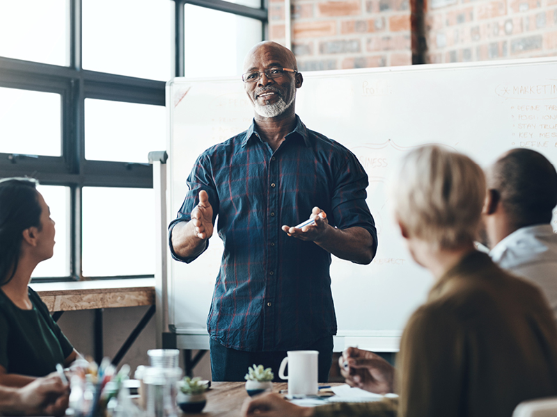 a man stands up at the front of a group of people sitting at a confrence table giving a speech