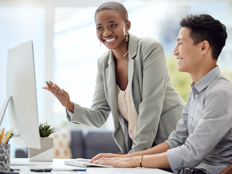 a woman stands next to a man sitting at a computer and poitns to computer screen while smiling