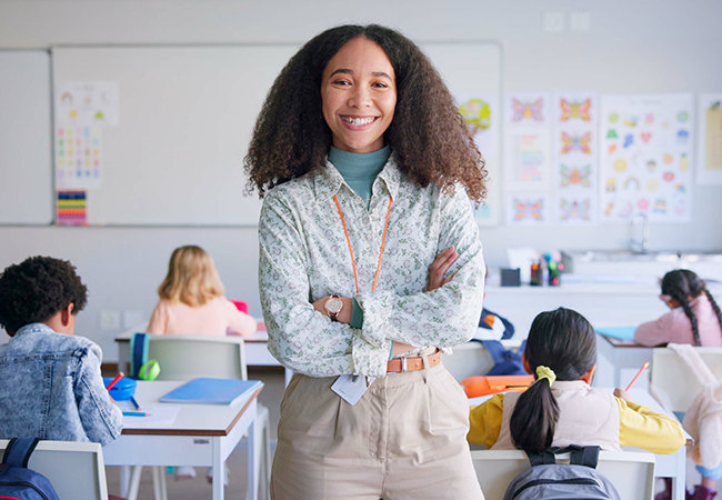 a professional woman stands in front of classroom with students behind her with arms crossed smiling at camera