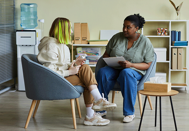a woman sits and listens to  student in chair accross from her with paper and pen in hand
