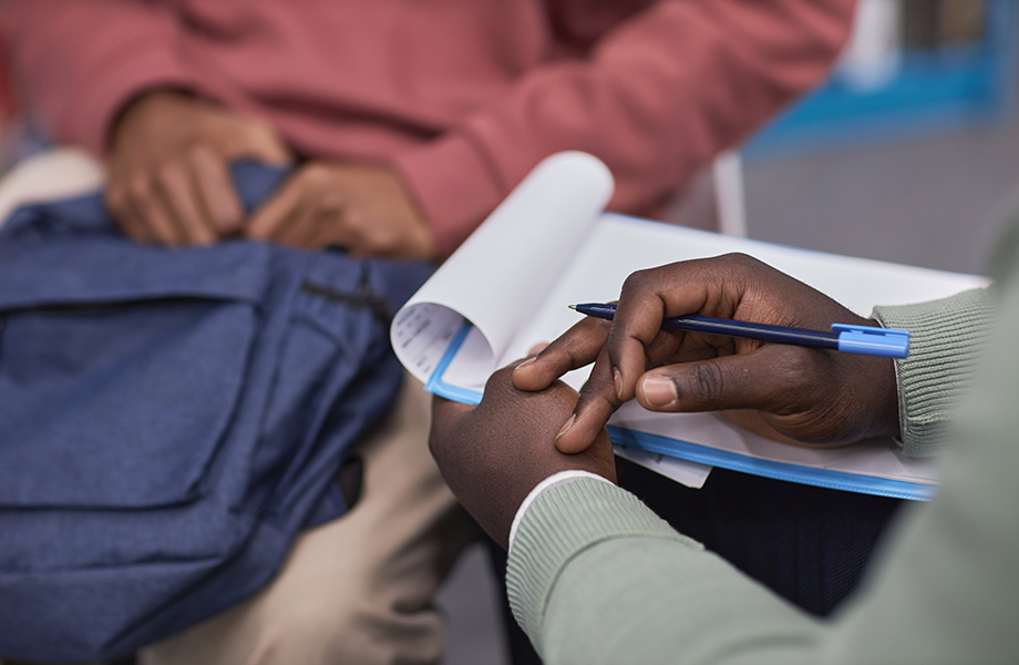 close up of hands holding papers and a pen sitting across from a boy with hands on a backpack