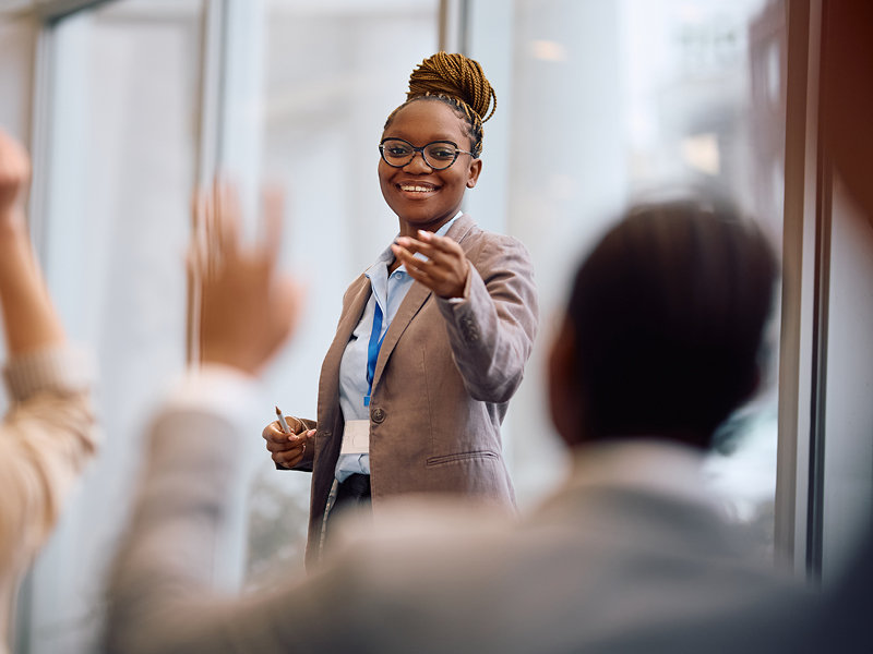 a woman stands in front of a confrence smiling and pointing at someone who is raising their hand