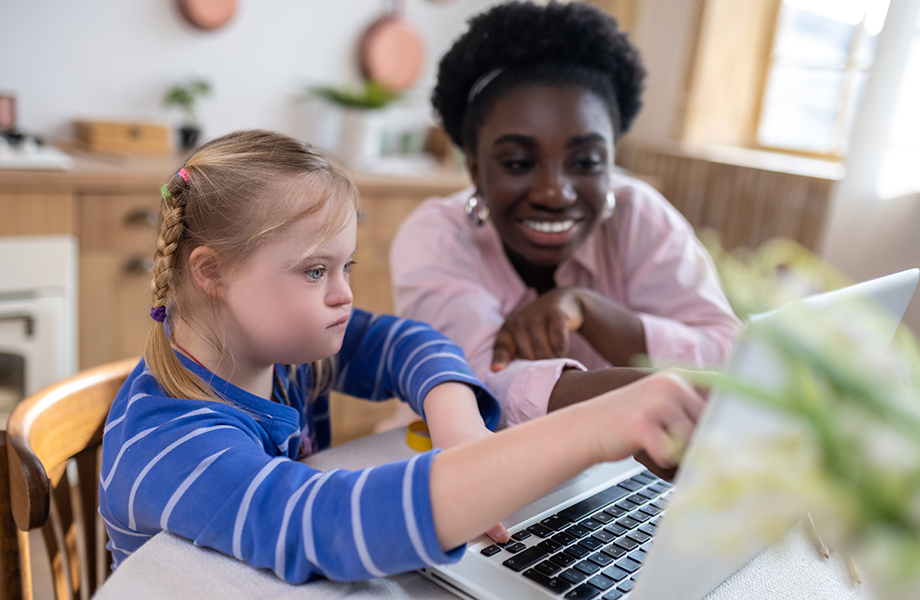 a young girl with down syndrome works on a computer with an instructor smiling behind pointing at screen