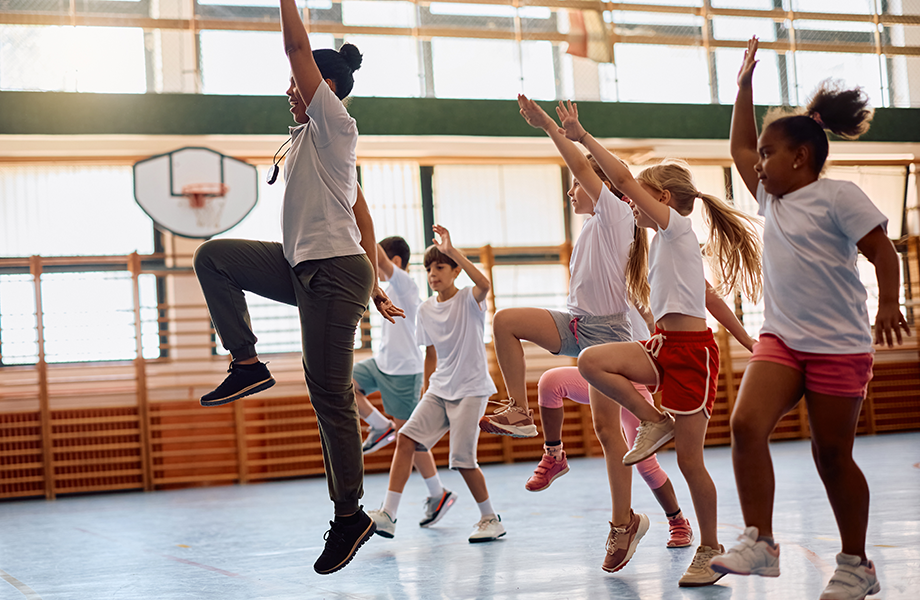a pe teacher stands in a gym with her class jumping up and down on one leg