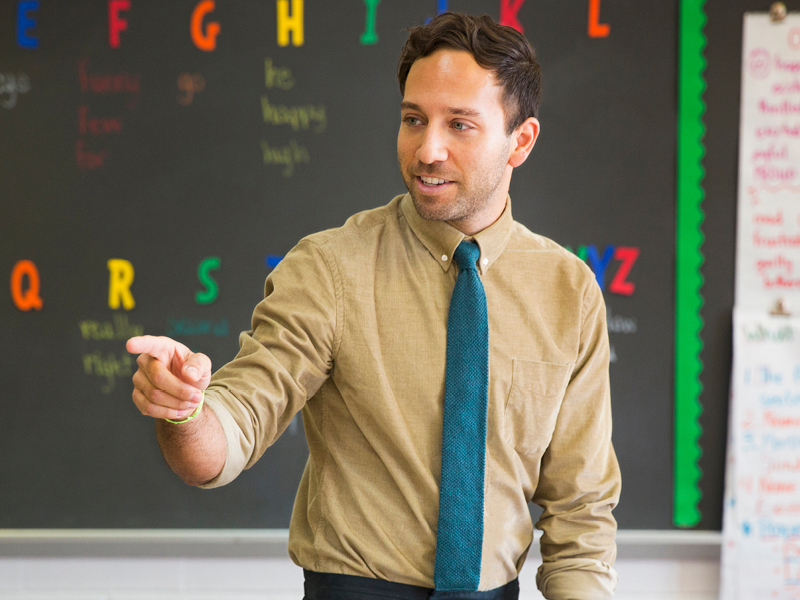 a male teacher stands in front of a blackboard pointing to a student in class