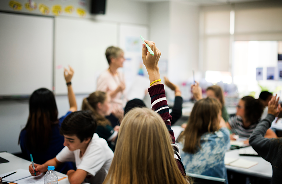 a wide shot of a classroom with the teacher out of focu and students raising their hands