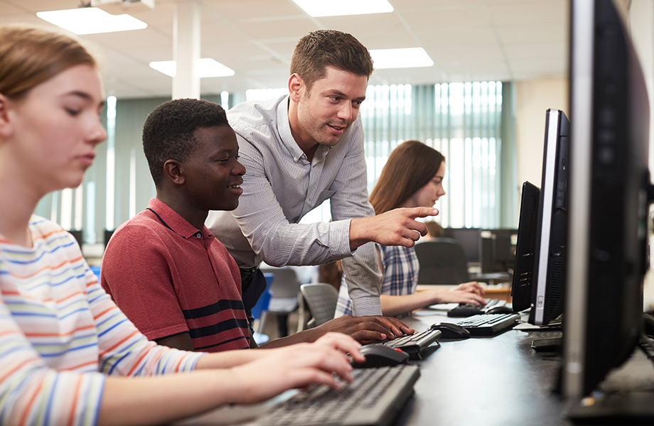 a teacher leans over a high school students sholder pointing at a computer screen