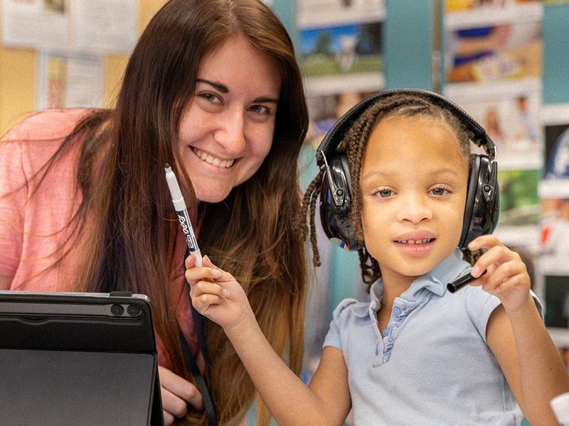 a teacher smiles with a student towards camera as the students wears headphones