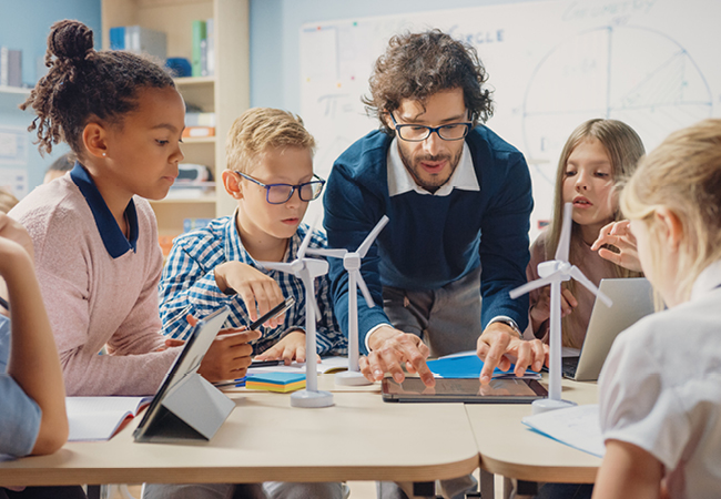 a male teacher leans over a table with elementary age students working on a tablet with a surpirsed look