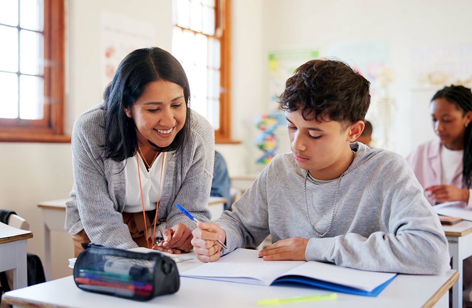 a woman teach leans over a student in class at his desk smiling
