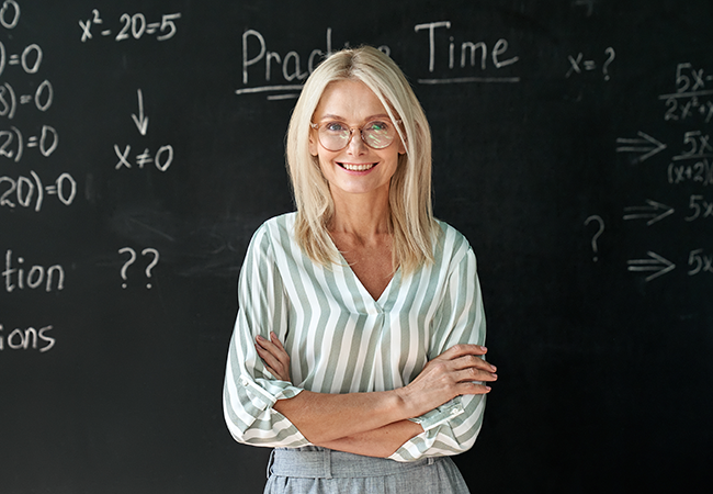 a teacher stands in front of a blackboard smiling at camera with math problems on board behid her