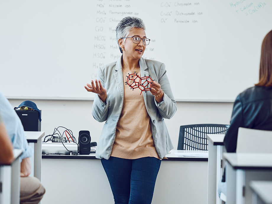 A science teacher stands at the front of a class holding a model of an atom