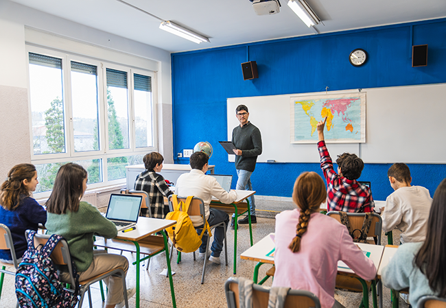 a teacher stands in front of a classroom with one student raising hand